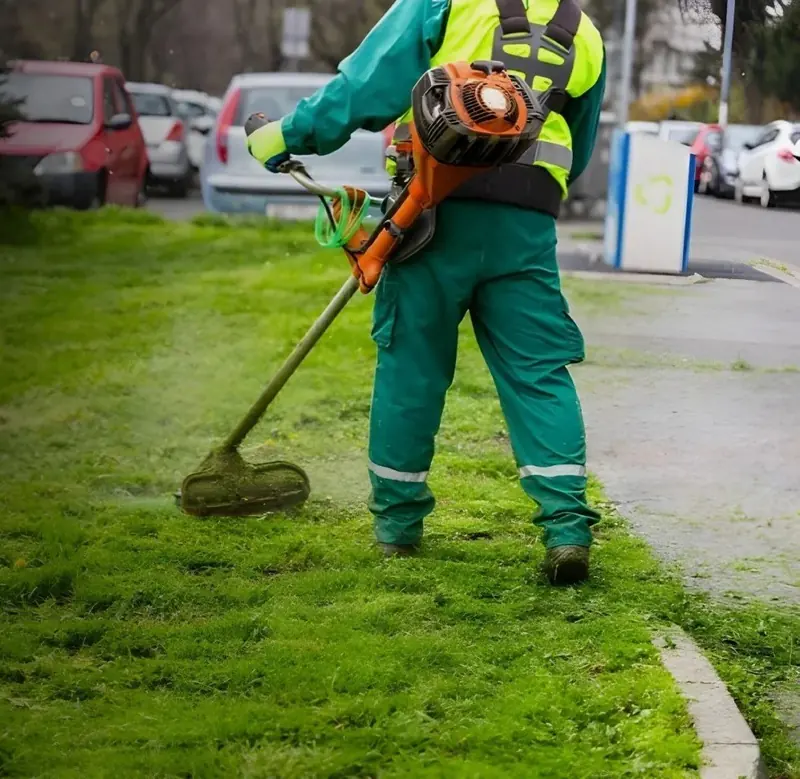 A worker using a string trimmer along the edge of a lawn beside a street.